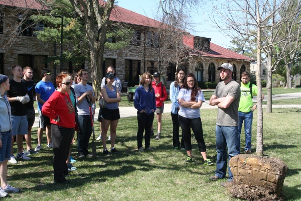 Group of about 15 people standing on lawn of Stauffer-Flint with a tree that is ready to be planted.