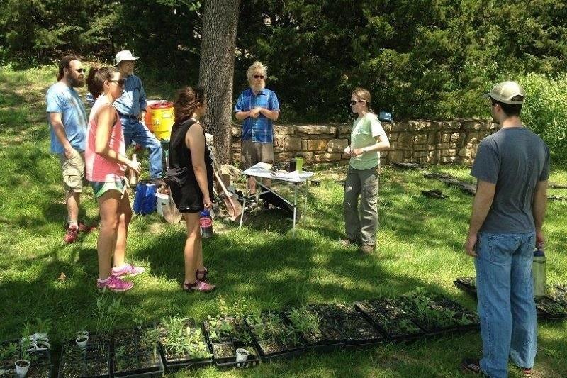 Group of people gathered under shade trees with trays of plant seedlings in the foreground ready to be planted.